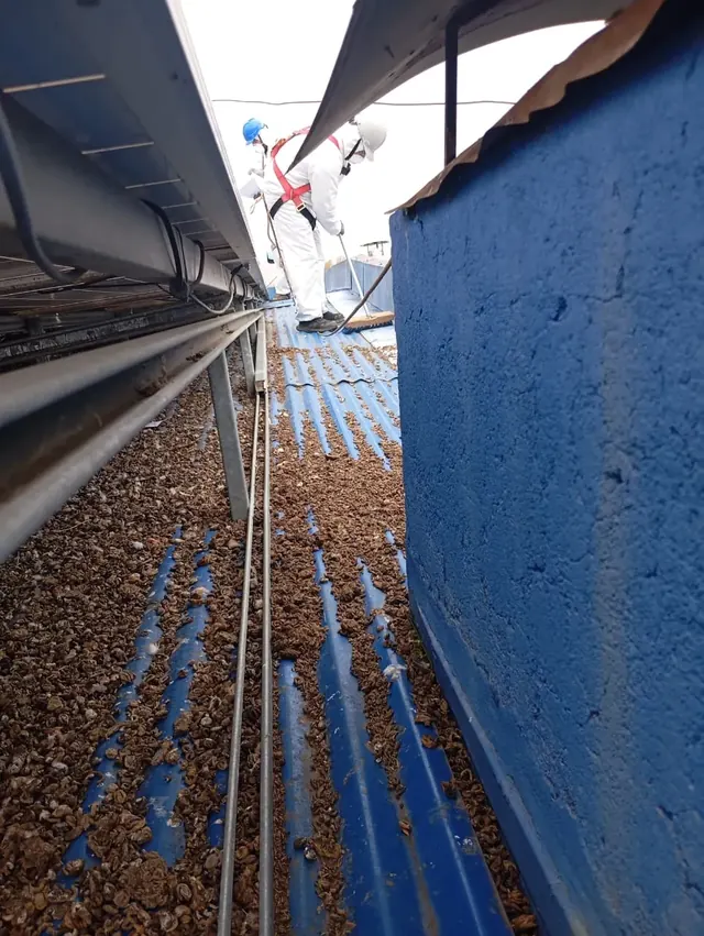 Personal técnico realizando limpieza bajo estructuras de paneles solares en techumbre industrial, siguiendo protocolos de seguridad. Personal con arnés realizando limpieza bajo paneles solares en cubierta azul.