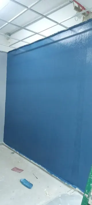 Aplicación de la identidad visual de la marca mediante recubrimientos de alta calidad. Muro con pintura fresca de color azul sólido bajo una estructura de cielo americano; se ven implementos de limpieza en el suelo de concreto.