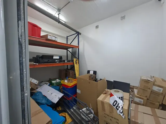 El punto de partida: un espacio de almacenamiento que se transformará en una sala de reuniones operativa. Bodega desordenada con estanterías metálicas naranjas, cajas de cartón acumuladas, herramientas y cables expuestos en un espacio de concreto.