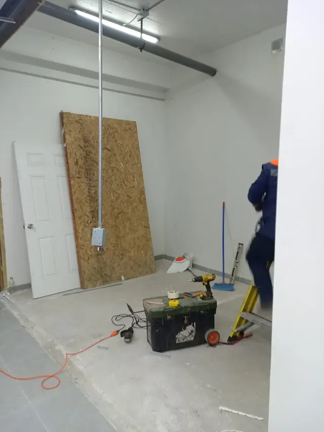 Mano de obra especializada ejecutando la canalización eléctrica y ajustes estructurales en tiempo real. Operario en escalera trabajando en instalaciones aéreas en una sala con paredes blancas, herramientas eléctricas y marcos de puertas apoyados.