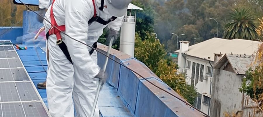 Trabajadores limpiando techumbre azul con arnés de seguridad.