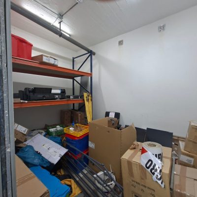 Bodega desordenada con estanterías metálicas naranjas, cajas de cartón acumuladas, herramientas y cables expuestos en un espacio de concreto.