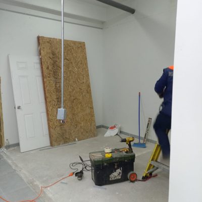 Operario en escalera trabajando en instalaciones aéreas en una sala con paredes blancas, herramientas eléctricas y marcos de puertas apoyados.