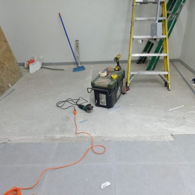 Primer plano de herramientas de construcción (lijadora, caja de herramientas, escalera) sobre un piso de concreto junto a un sector de porcelanato gris terminado.
