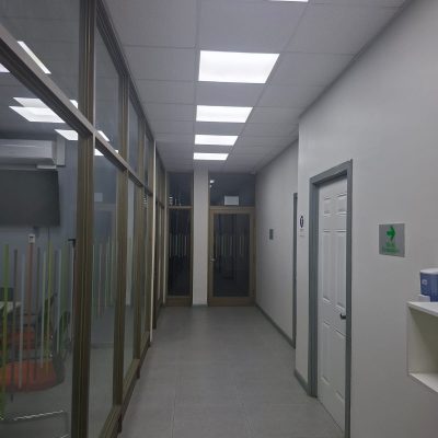 Pasillo interior de oficina con piso de porcelanato gris, mamparas de cristal con marcos metálicos a la izquierda y cielo falso con iluminación LED integrada.
