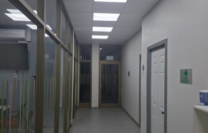 Pasillo interior de oficina con piso de porcelanato gris, mamparas de cristal con marcos metálicos a la izquierda y cielo falso con iluminación LED integrada.