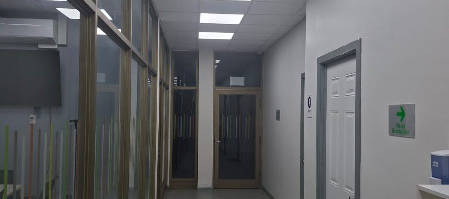 Pasillo interior de oficina con piso de porcelanato gris, mamparas de cristal con marcos metálicos a la izquierda y cielo falso con iluminación LED integrada.